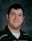 A smiling young man with short dark hair, wearing a black and white striped shirt, against a dark background.