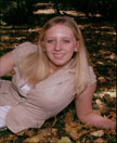 A young woman with long blonde hair is lying on a bed of autumn leaves, smiling at the camera while wearing a light-colored top and a beige vest.