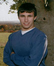 A young man stands with his arms crossed in front of a tree, wearing a blue sweater, with a scenic landscape in the background.