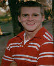 A young man with short hair smiles while wearing a red and white striped shirt, standing outdoors near a tree.