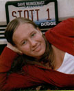 A young woman with long hair is lying on the ground, smiling, in front of a vehicle with a license plate that reads 'STOTT 1'.