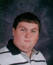 A young man with short brown hair, wearing a black and white striped polo shirt, poses for a school portrait against a dark background.