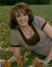 A smiling woman with curly hair is lying on a grassy area covered with autumn leaves, wearing a striped top.
