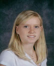 A young girl with long blonde hair, wearing a white shirt, smiles for a school portrait against a dark background.