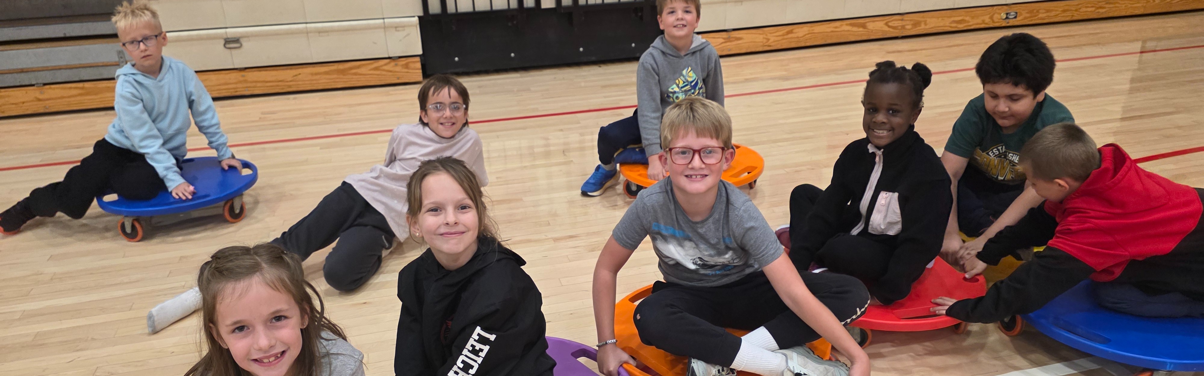 Students sitting and smiling on colorful scooter boards in the school gym.