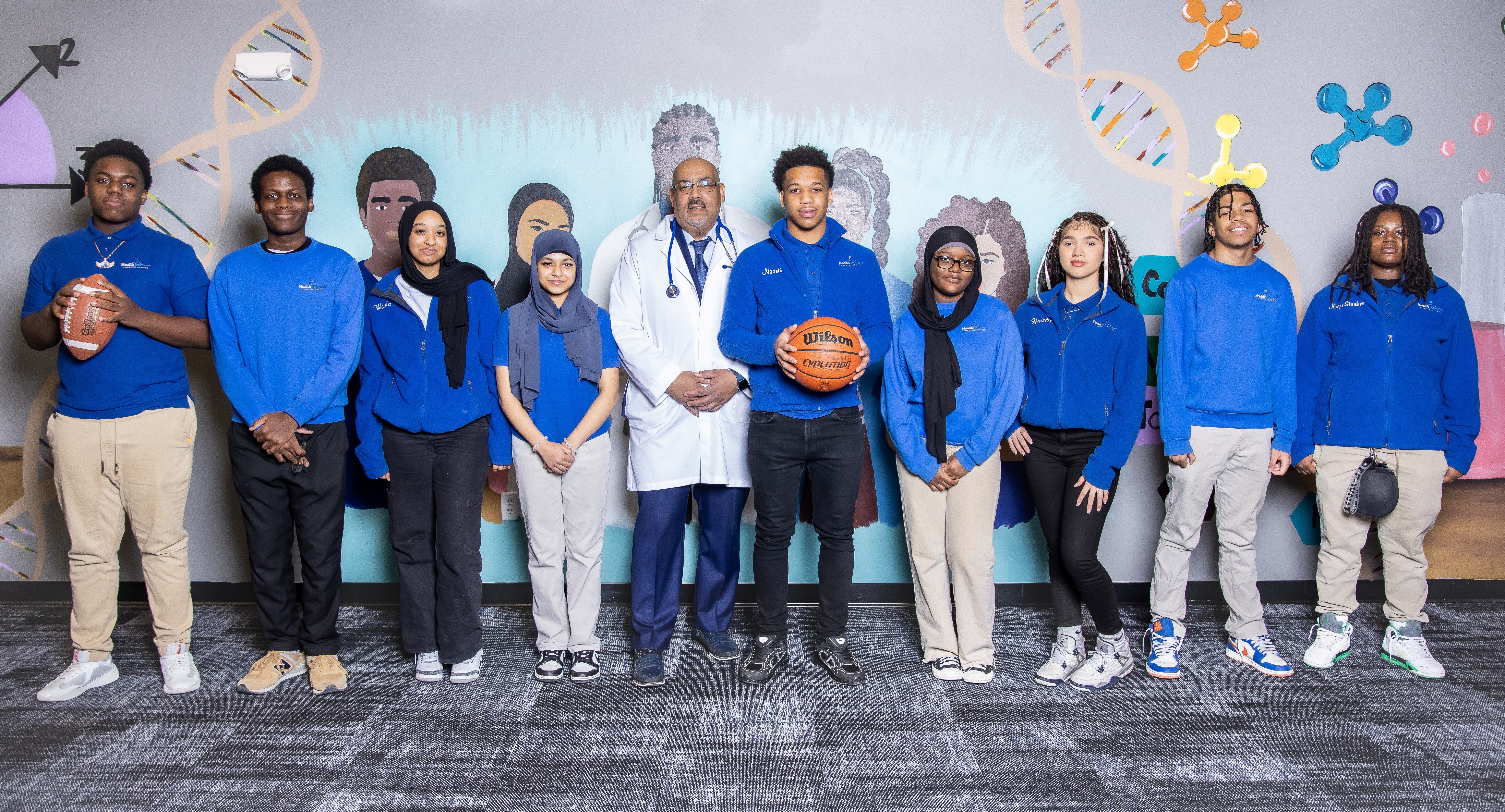 A picture of students standing next to a wall with a science theme. From right to left there is a boy holding a football, another boy standing next to him, two girls in hijabs, the Head of School Mr. Baxter in a doctor's coat, a boy holding a basketball, another girl wearing a hijab, and one girl and two boys.