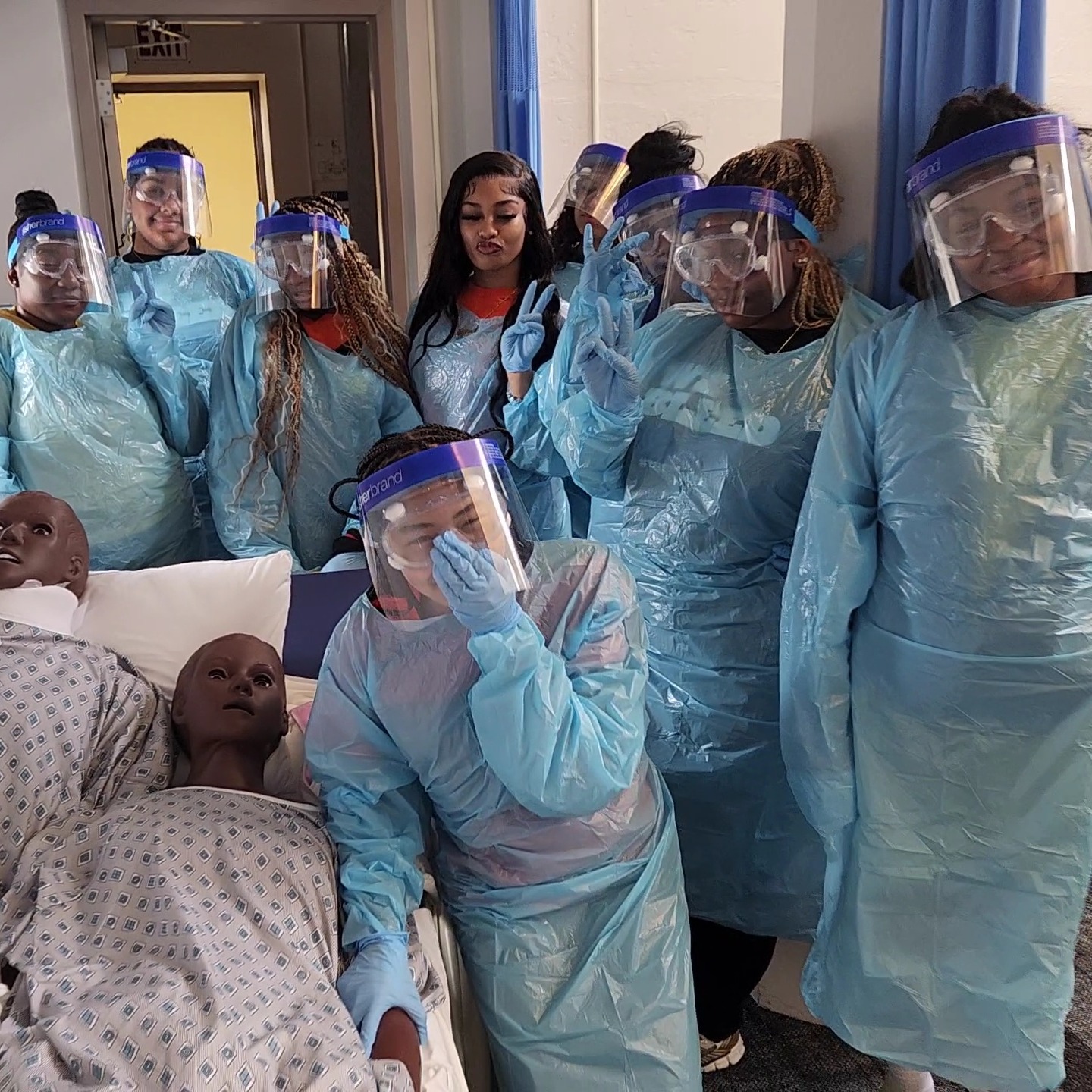 High school students wearing blue medical gowns, face shields, and masks gather around medical training mannequins in a clinical lab setting.