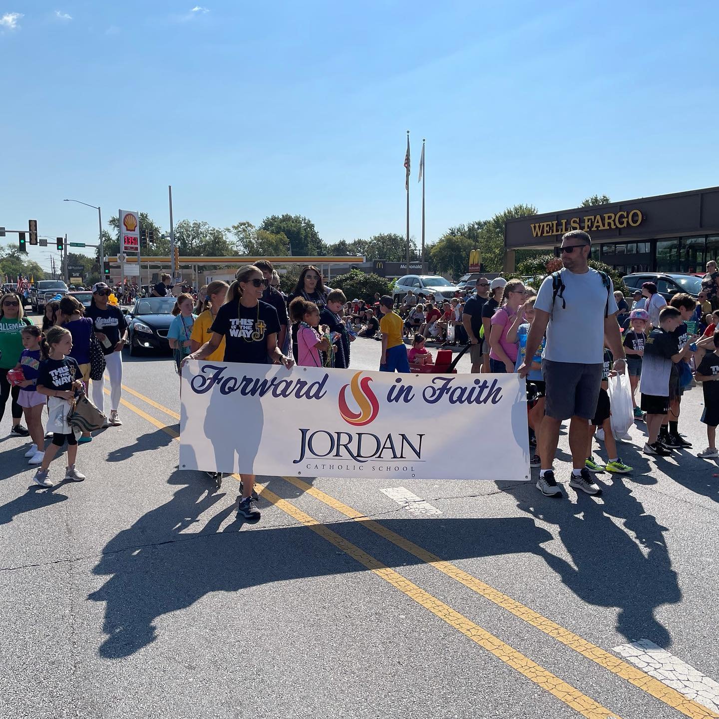A group of people, including children and adults, walking in a parade while holding a large banner that reads 'Forward in Faith' with the name 'Jordan' underneath, set against a sunny day with a street and buildings in the background.