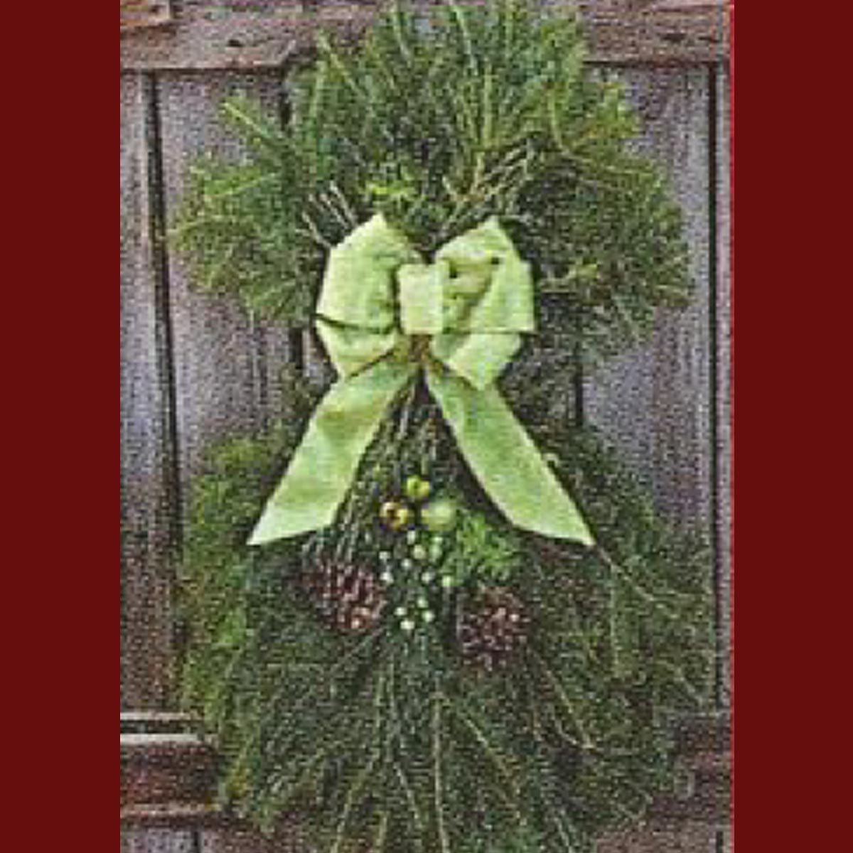 A decorative green wreath adorned with a light green bow and small pine cones, hanging against a wooden fence.