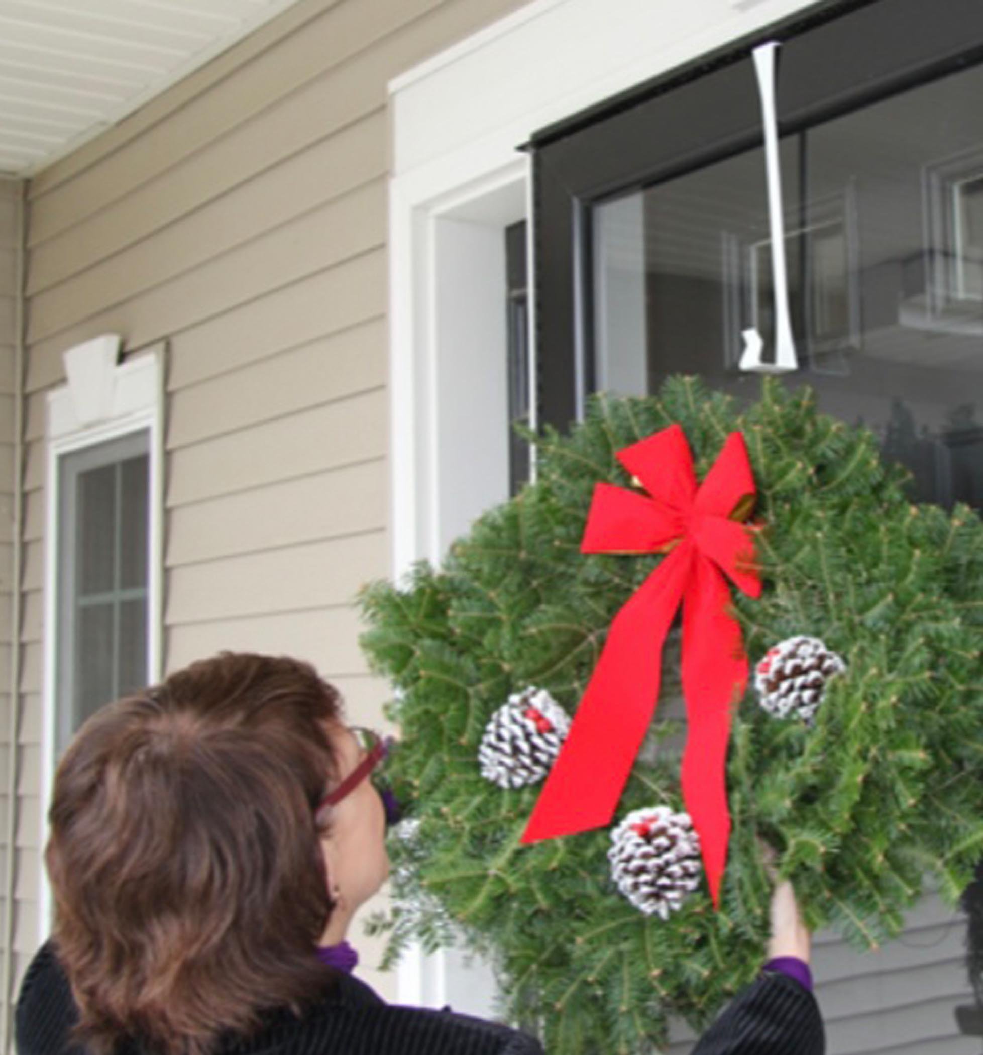 A person is hanging a festive green wreath adorned with a red bow and pine cones on a front door.