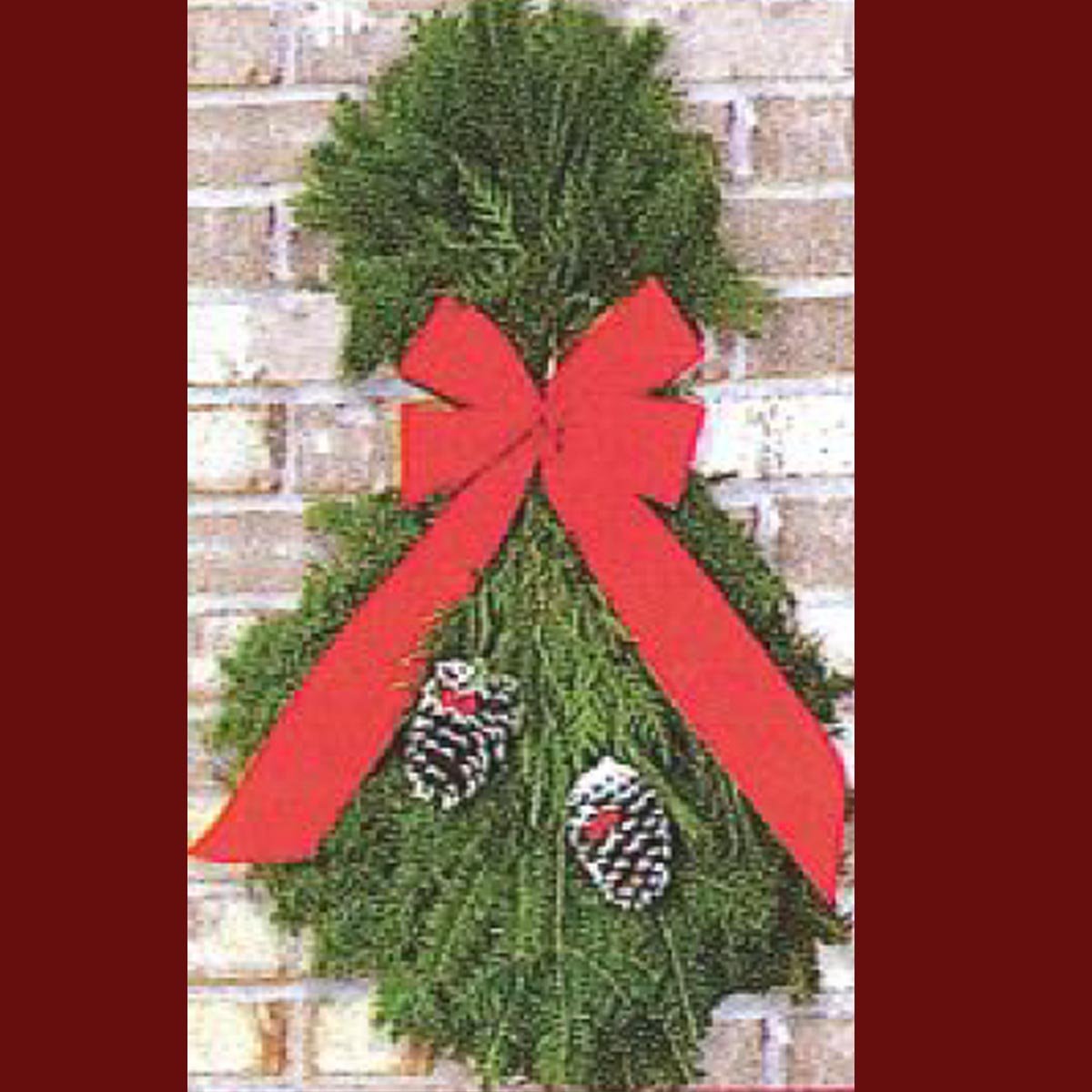 A decorative Christmas wreath made of evergreen branches, featuring a large red bow and two pine cones, hanging against a brick wall.