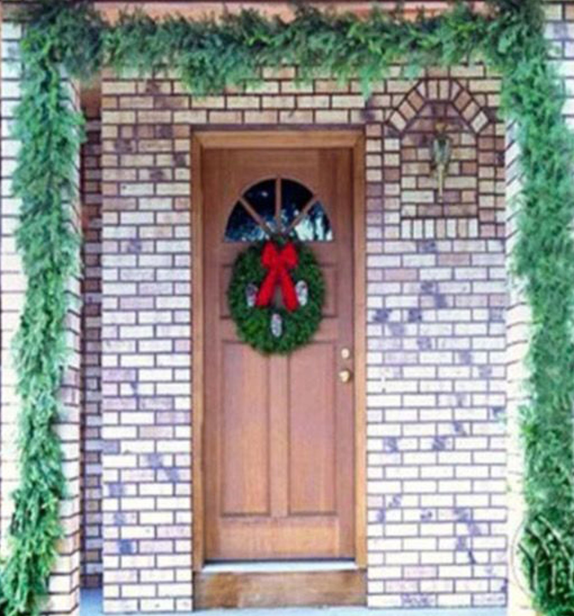 A decorated front door featuring a green wreath with a red bow, surrounded by garlands of greenery, set against a brick wall.