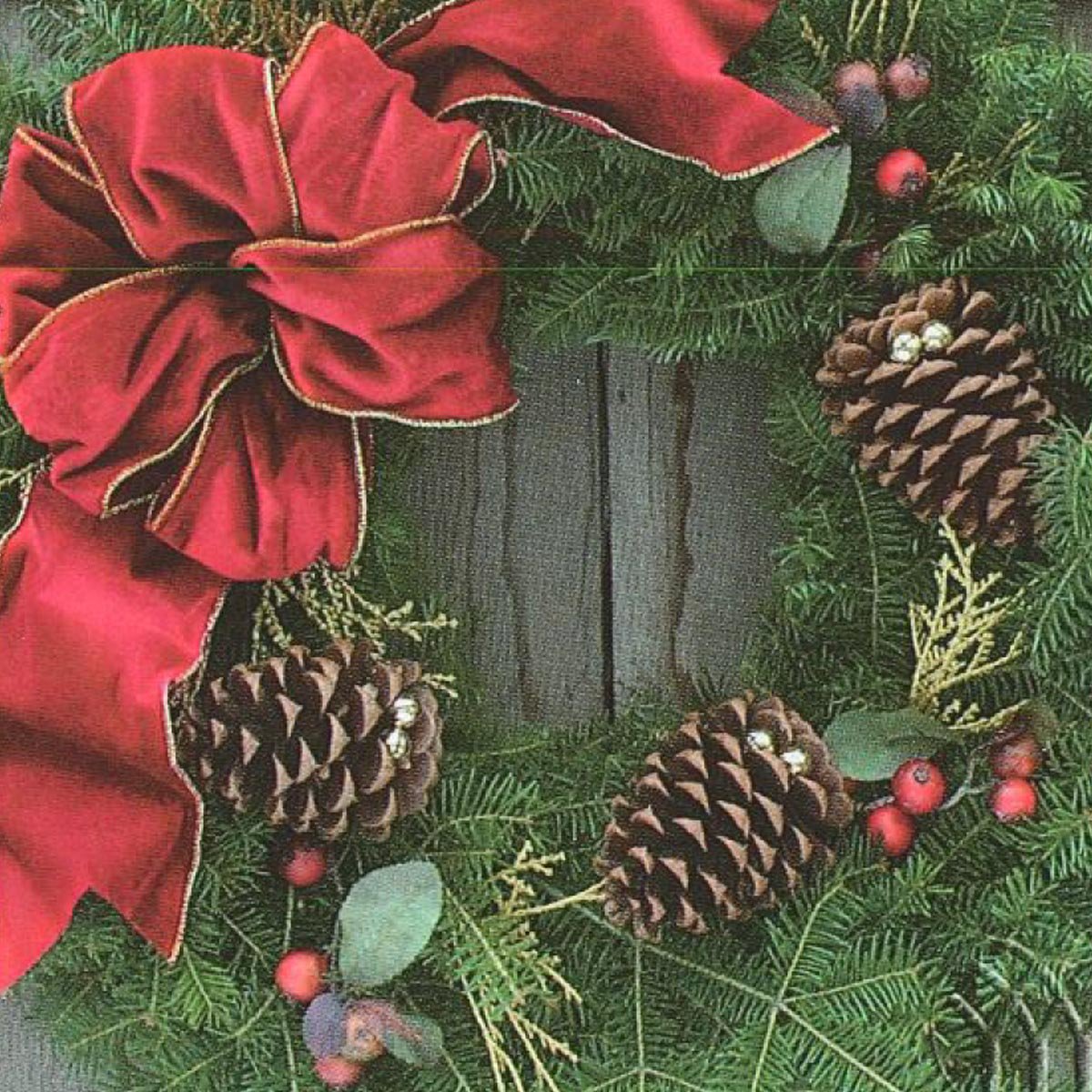 A festive holiday wreath adorned with a large red bow, pinecones, and greenery, set against a wooden background.