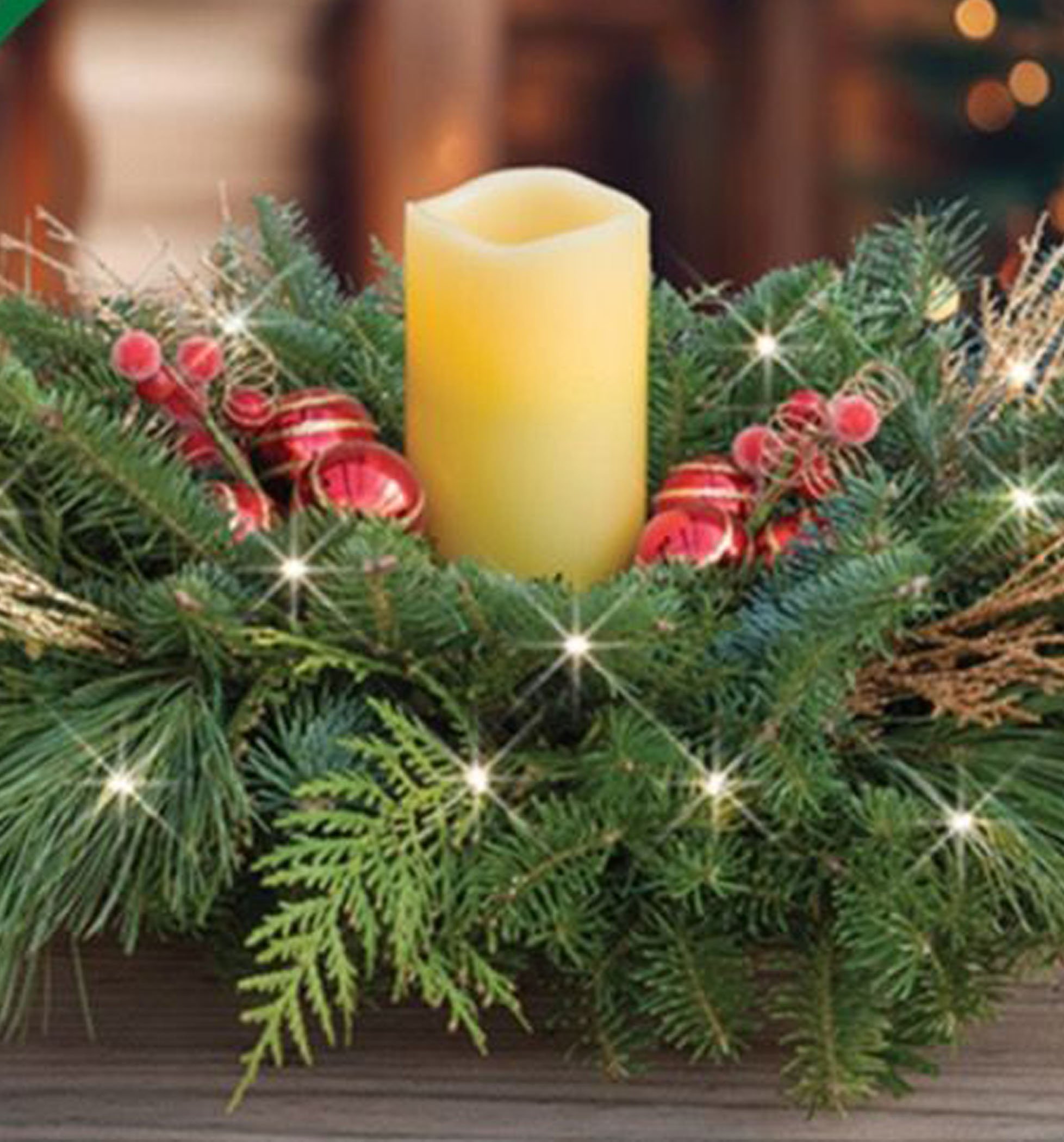 A decorative arrangement featuring a yellow candle surrounded by green pine branches, red ornaments, and twinkling lights, evoking a festive holiday atmosphere.