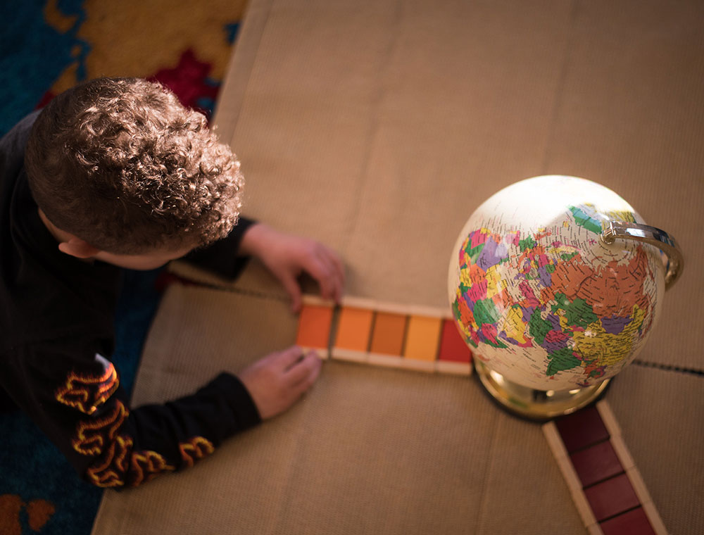 A child with curly hair is arranging colored blocks next to a globe on a carpeted surface, focusing on the task at hand.