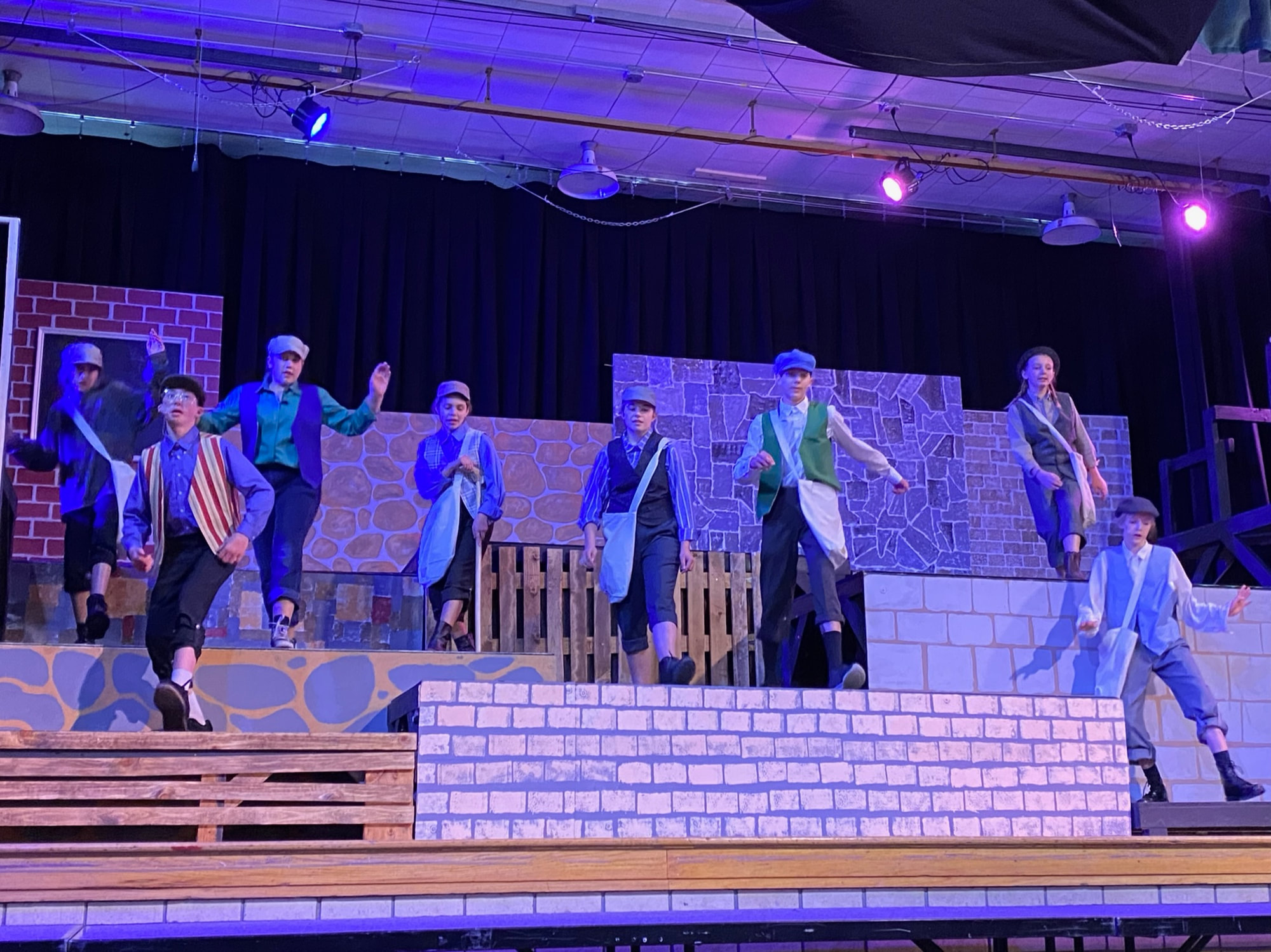A group of performers in colorful costumes are dancing on a stage set designed with brick walls and wooden structures, illuminated by purple stage lights.