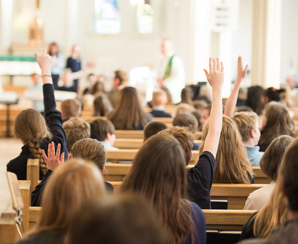 A group of children in a church setting, with many raising their hands, indicating participation or eagerness to answer questions.