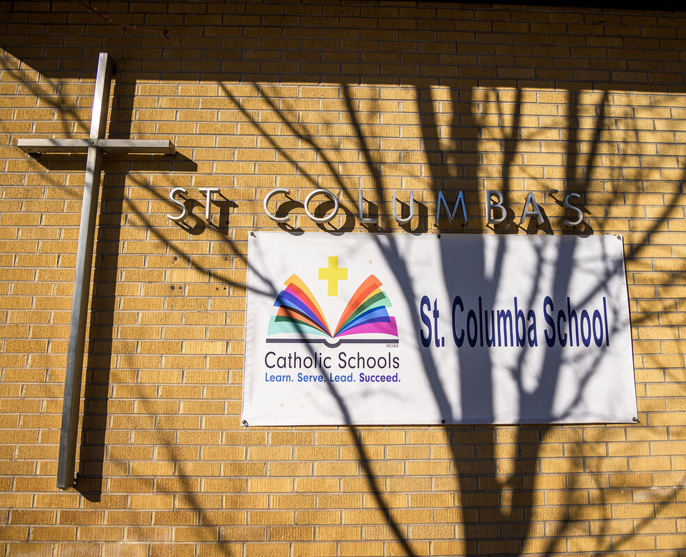 The exterior of St. Columba School features a large cross and a banner displaying the school's name and motto, with tree shadows cast on the brick wall.