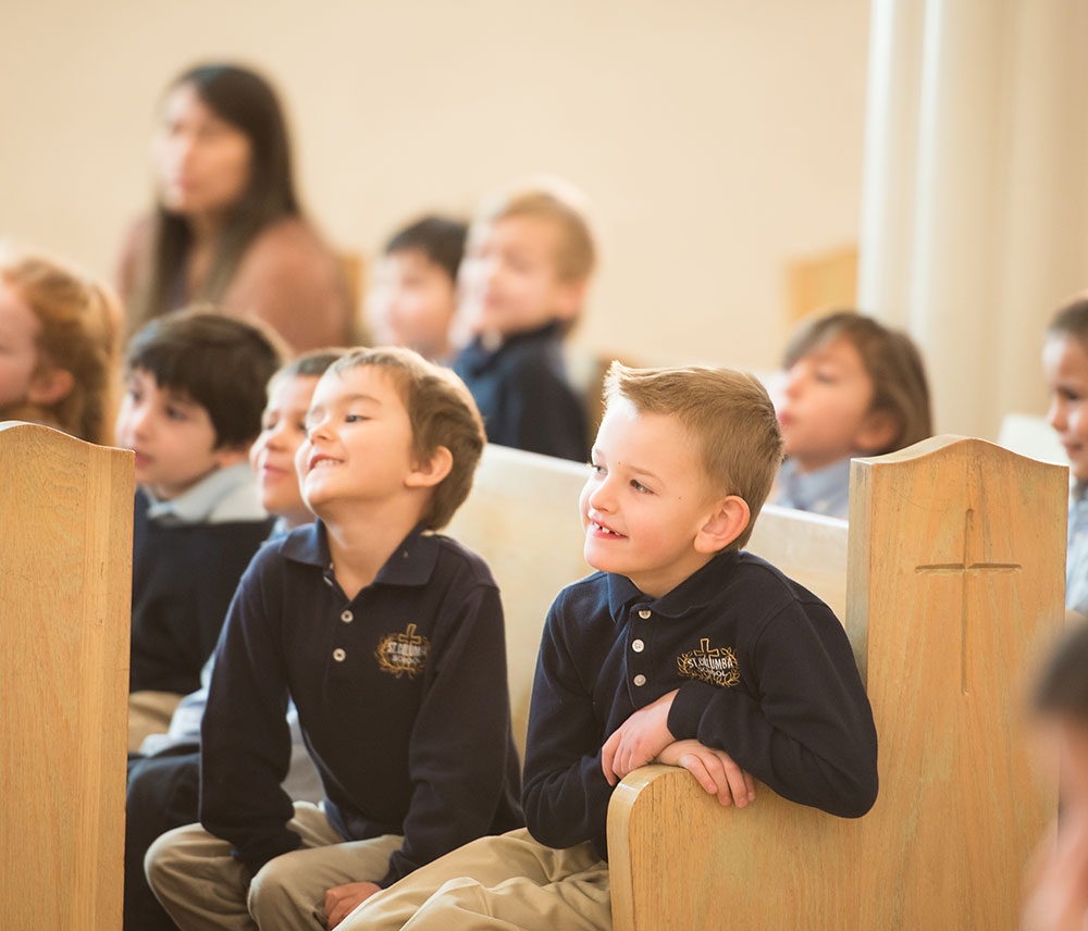A group of children sitting in a church, some smiling and engaged, while others appear attentive to the front.