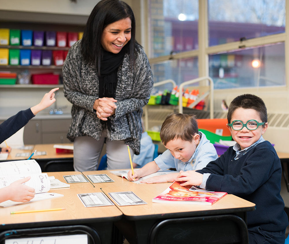 A teacher interacts with students at a classroom table, where two boys are engaged in activities while another student raises a hand.