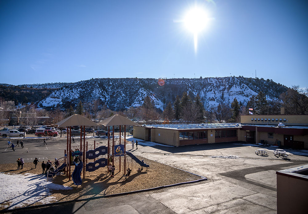 A sunny winter day at a school playground, with children playing and snow-covered hills in the background.
