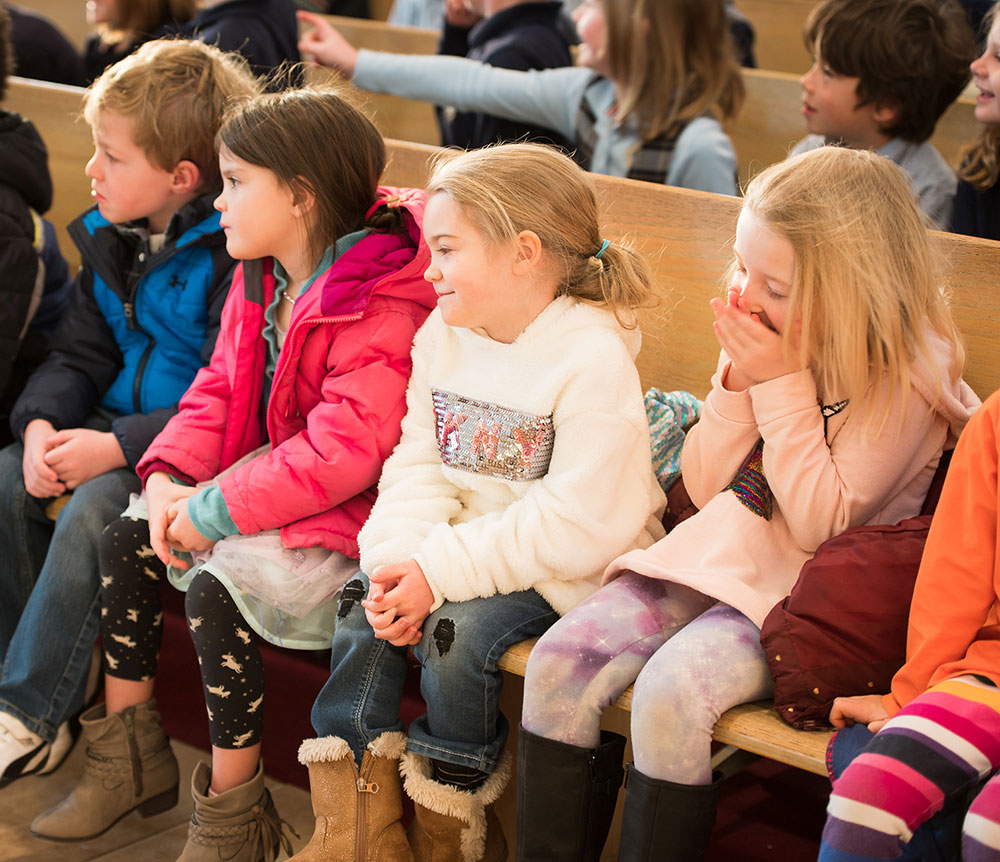 A group of children sitting on wooden benches, some wearing colorful jackets and boots, with expressions ranging from attentive to playful.