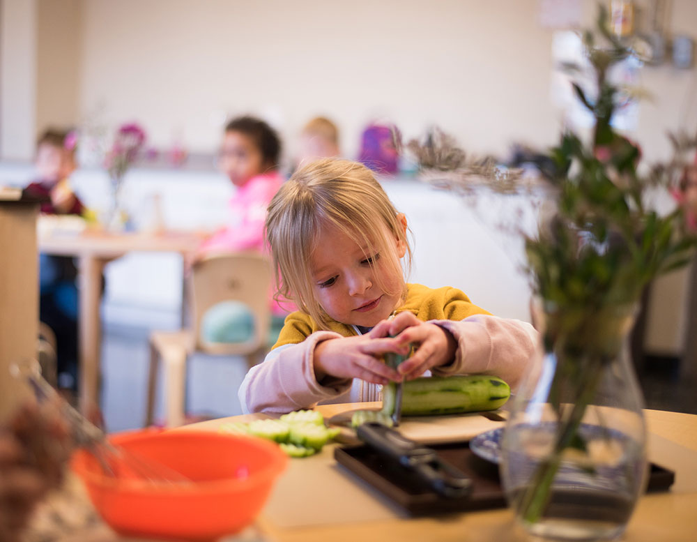 A young child with blonde hair is focused on cutting vegetables at a table, surrounded by other children in a classroom setting.