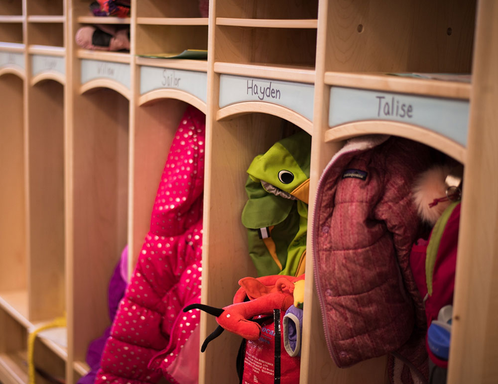 A wooden cubby storage unit displaying colorful children's jackets and costumes, with labels for each child's name above the compartments.