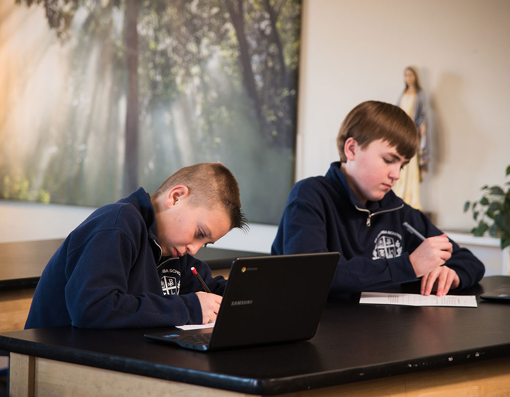 Two boys are seated at a table in a classroom, one is focused on writing in a notebook while the other is using a laptop, with a serene background of trees and soft lighting.