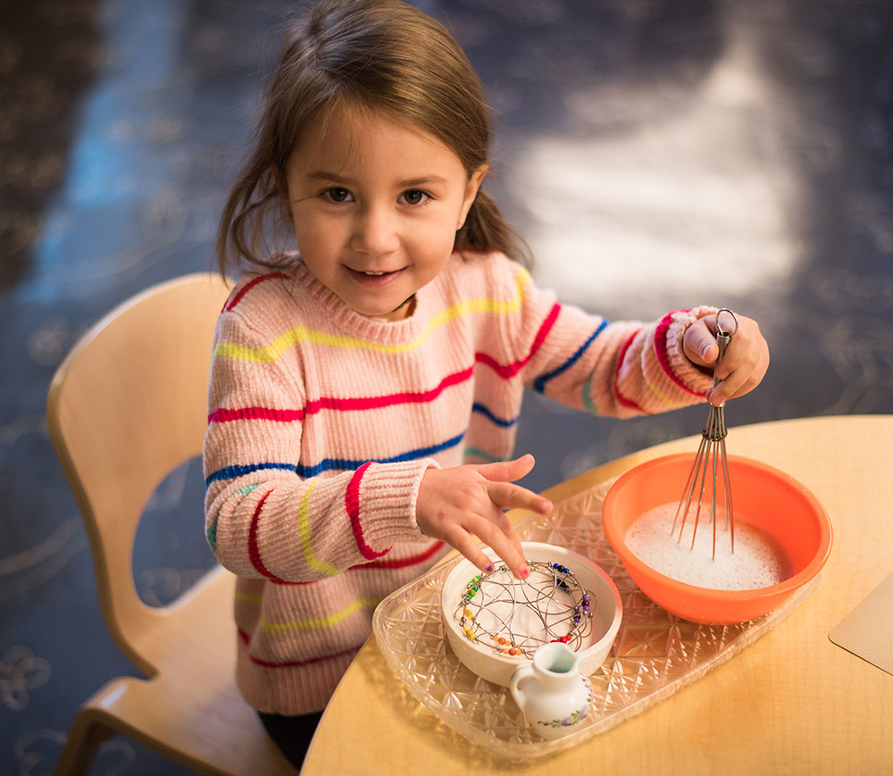 A young girl with brown hair, wearing a striped sweater, smiles while holding a whisk over a bowl of white sugar, with a small pitcher and a plate of colorful sprinkles on the table in front of her.