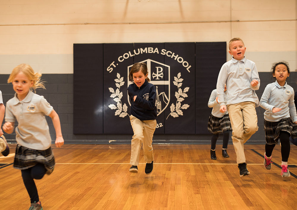 A group of children in school uniforms are running energetically across a gymnasium floor, with a school emblem displayed on the wall behind them.