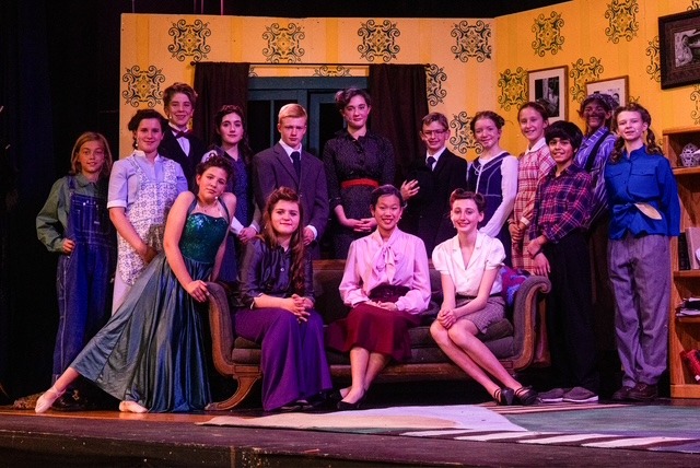 A group of performers in colorful costumes are dancing on a stage set designed with brick walls and wooden structures, illuminated by purple stage lights.