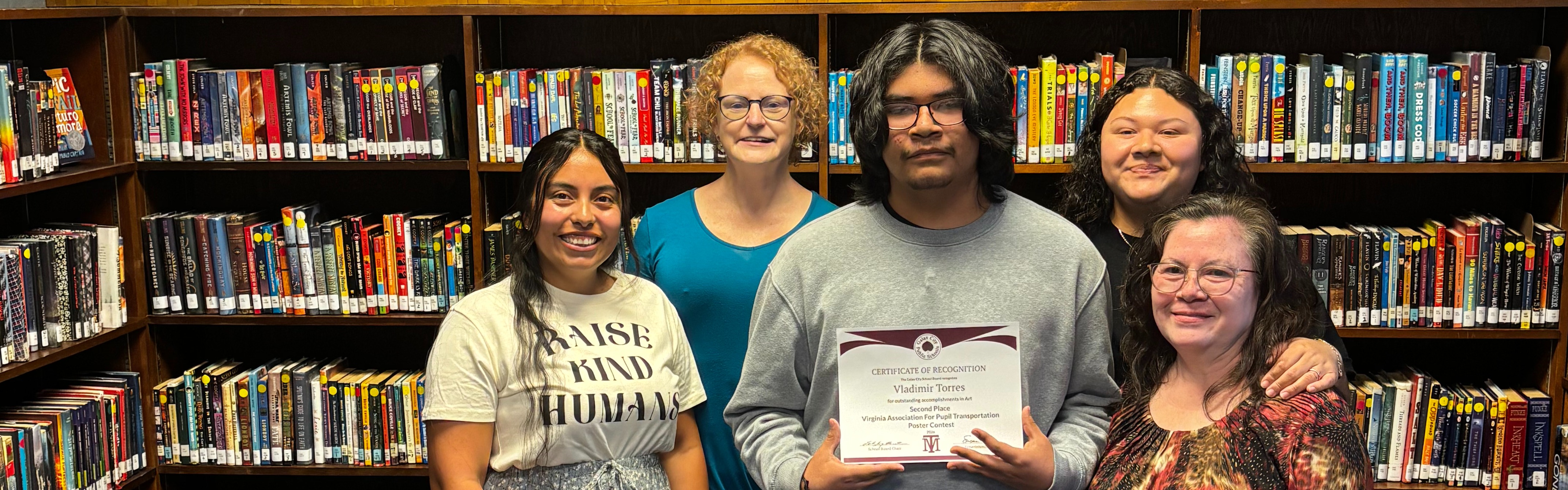 Five people stand in a library, smiling. One holds a certificate. Shelves with books behind them. "GALAX" on the wall.