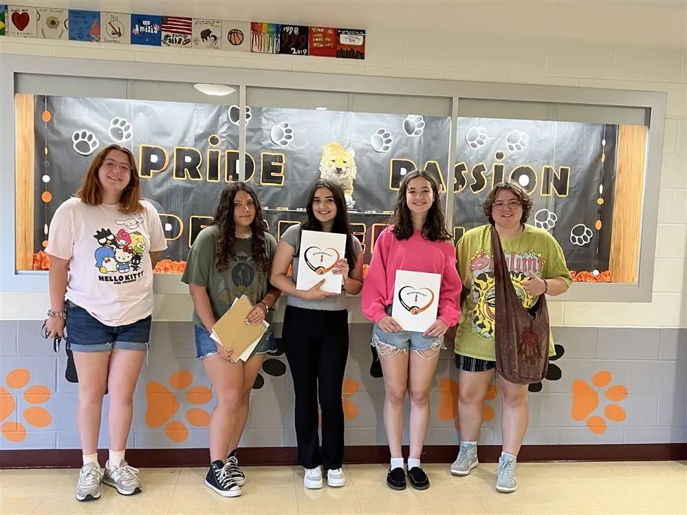 A group of five students stands in front of a school display featuring the words "Pride Passion" on a black backdrop, each holding art.