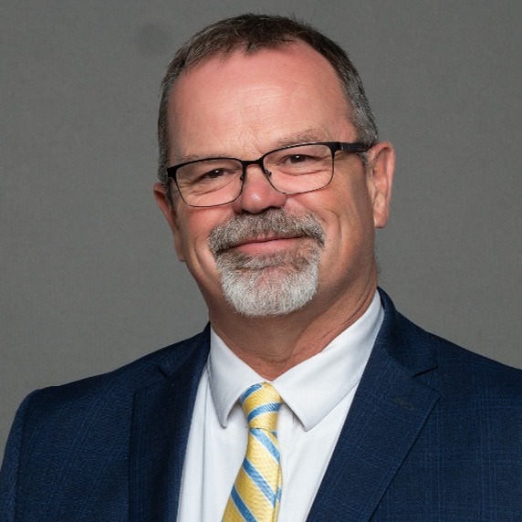 A man in a suit and tie poses for a professional headshot against a blue background.