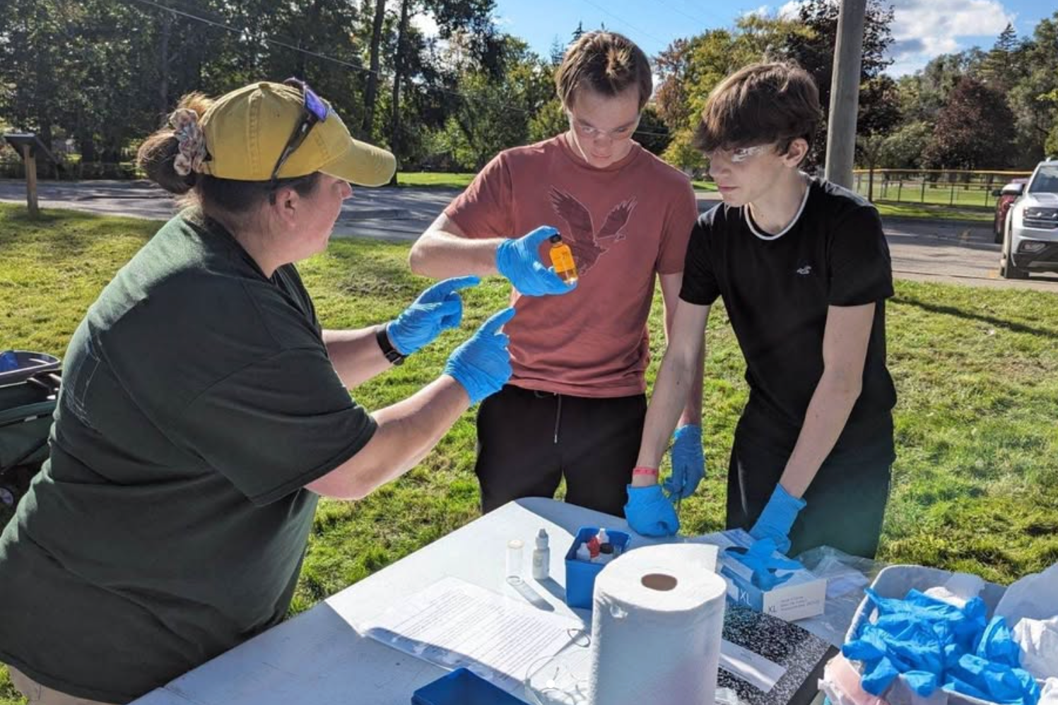 Students study a bottle with blue latex gloves on. Teacher is explaining something to them outside. 