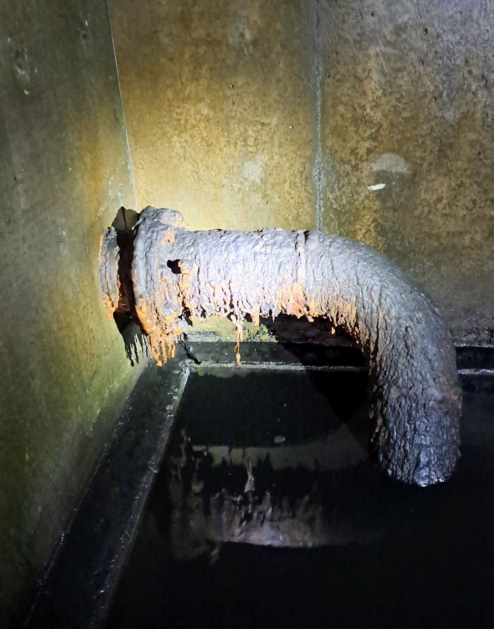 A rusty pipe extends from a corner of a concrete wall into a dark, wet area.