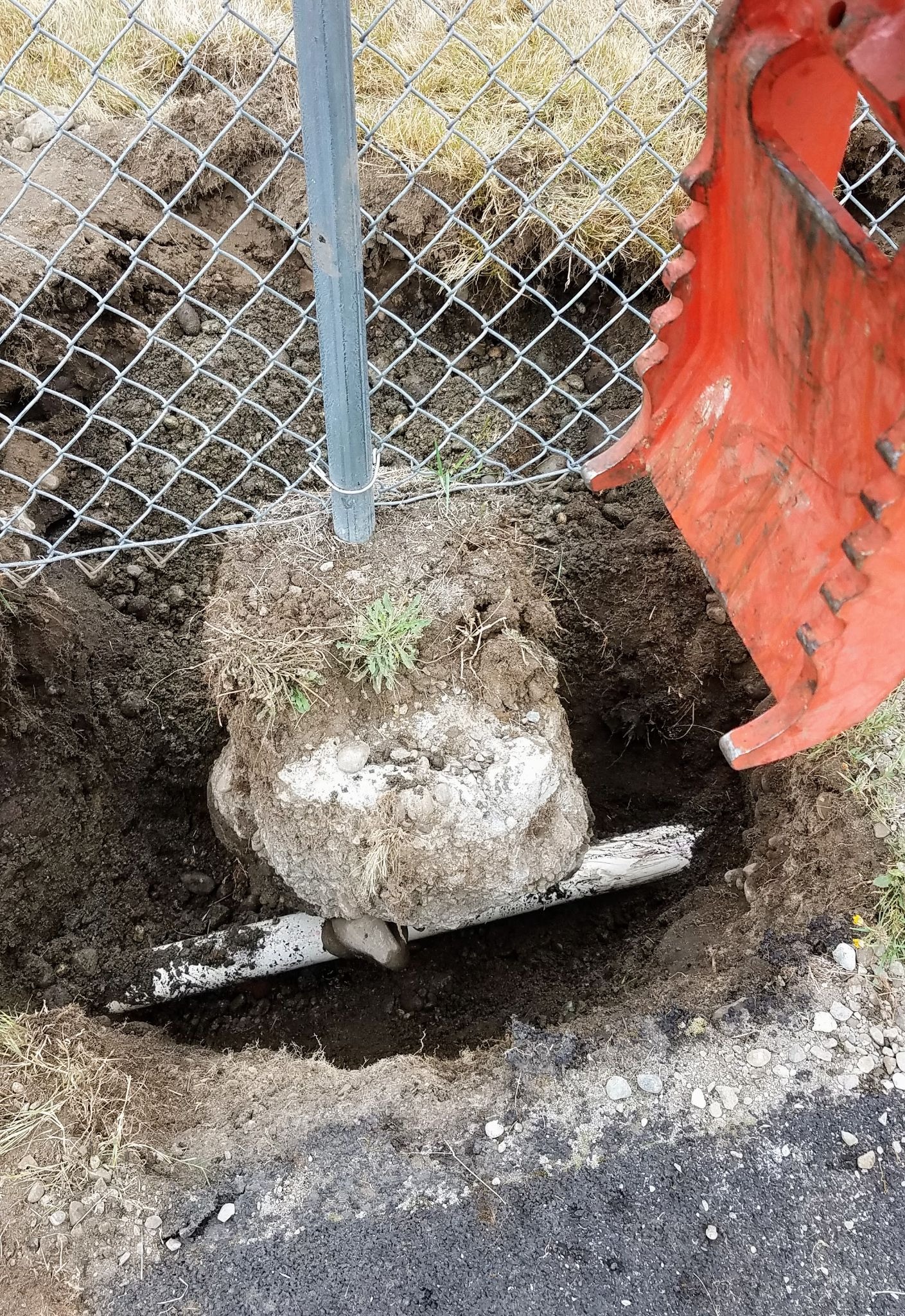 A red construction machine lifts a pole from the ground, revealing a partially exposed rock. The area is fenced.