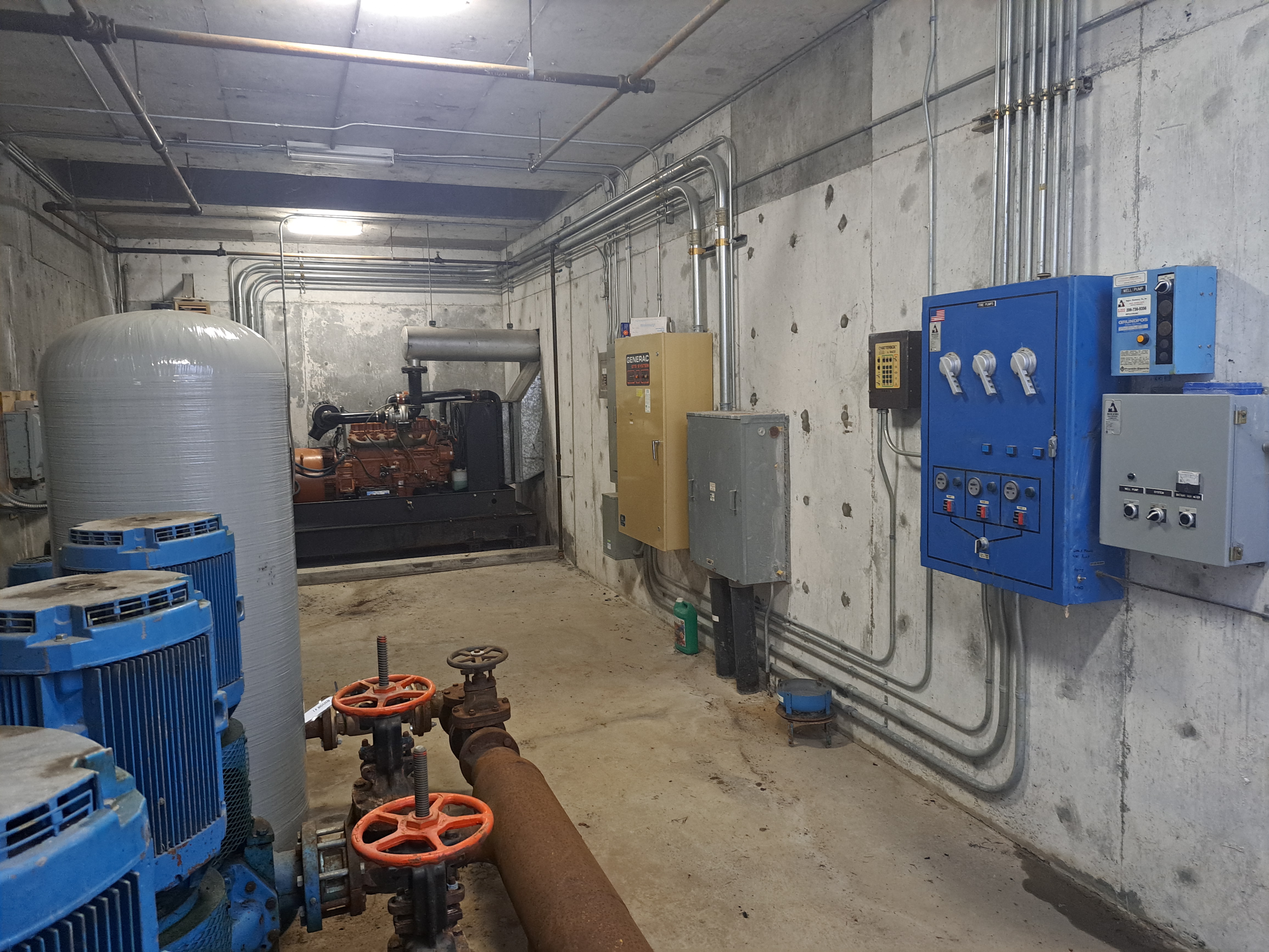 An industrial room with machinery. Large tanks and pipes are on the floor. There are control panels on the wall.