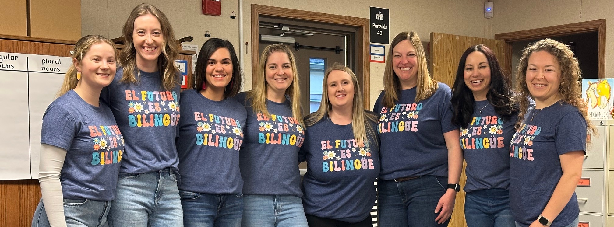 Seven women in blue shirts with colorful designs and jeans stand in a room with a white board and cabinets.