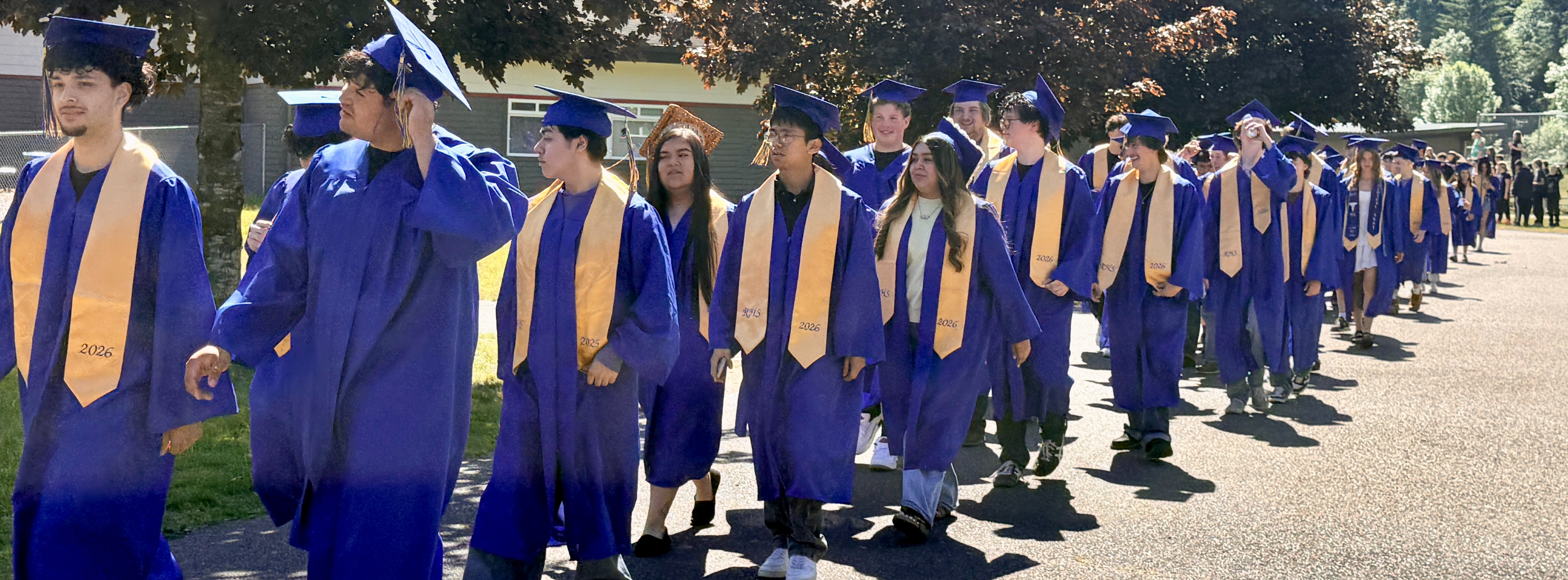 Students in blue graduation gowns and caps walk in a line on a sidewalk, lined with trees.