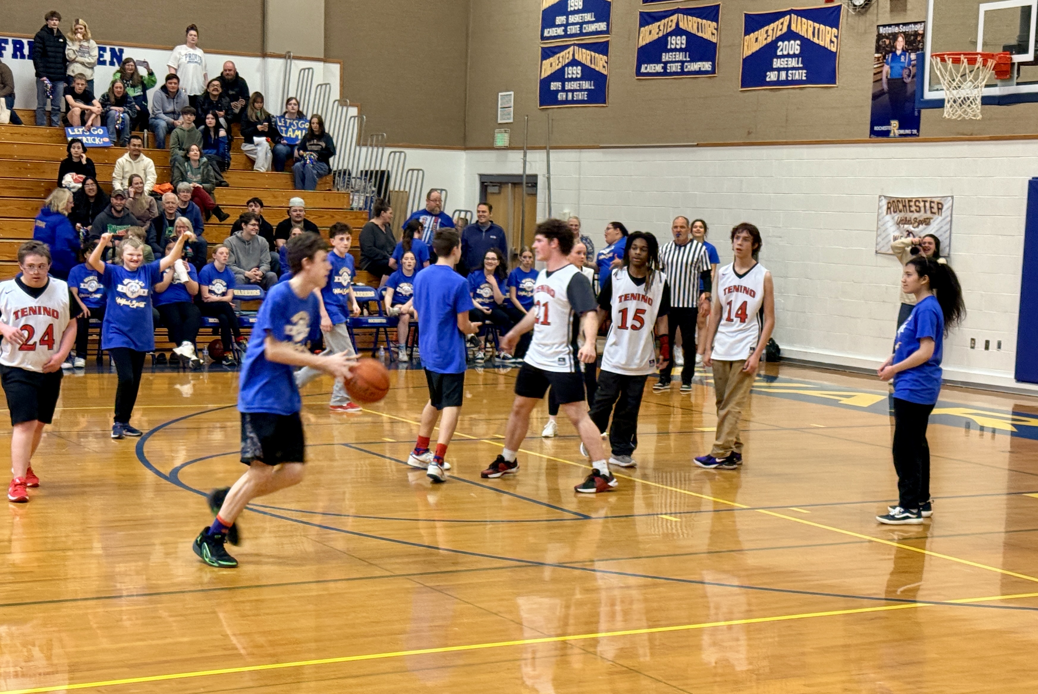 A basketball game in a gym. Players wear numbered jerseys. A player dribbles the ball, and others move around. Spectators are on the bleachers.