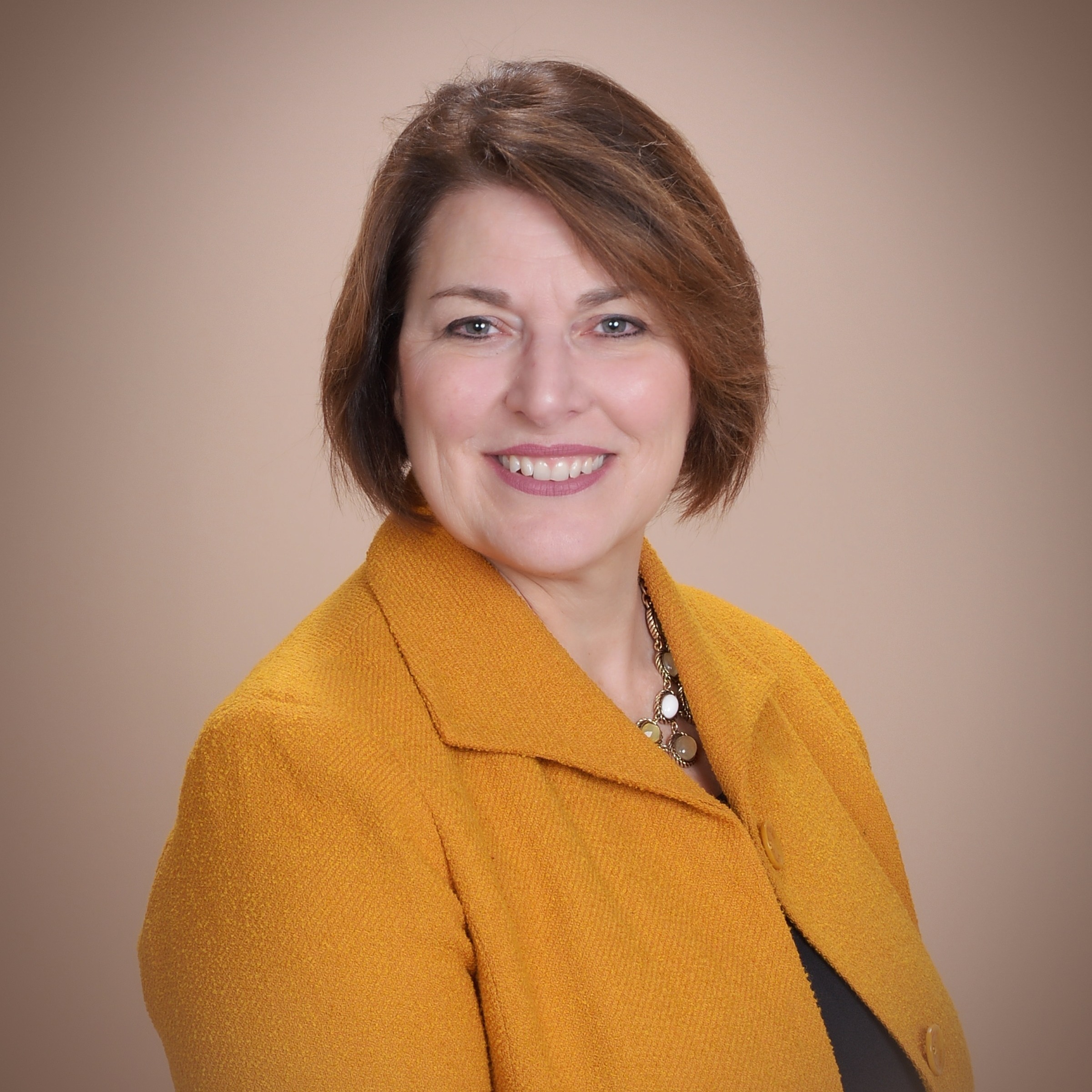 A woman with short hair smiles, wearing a yellow blazer over a black top. She has a necklace. The background is beige.