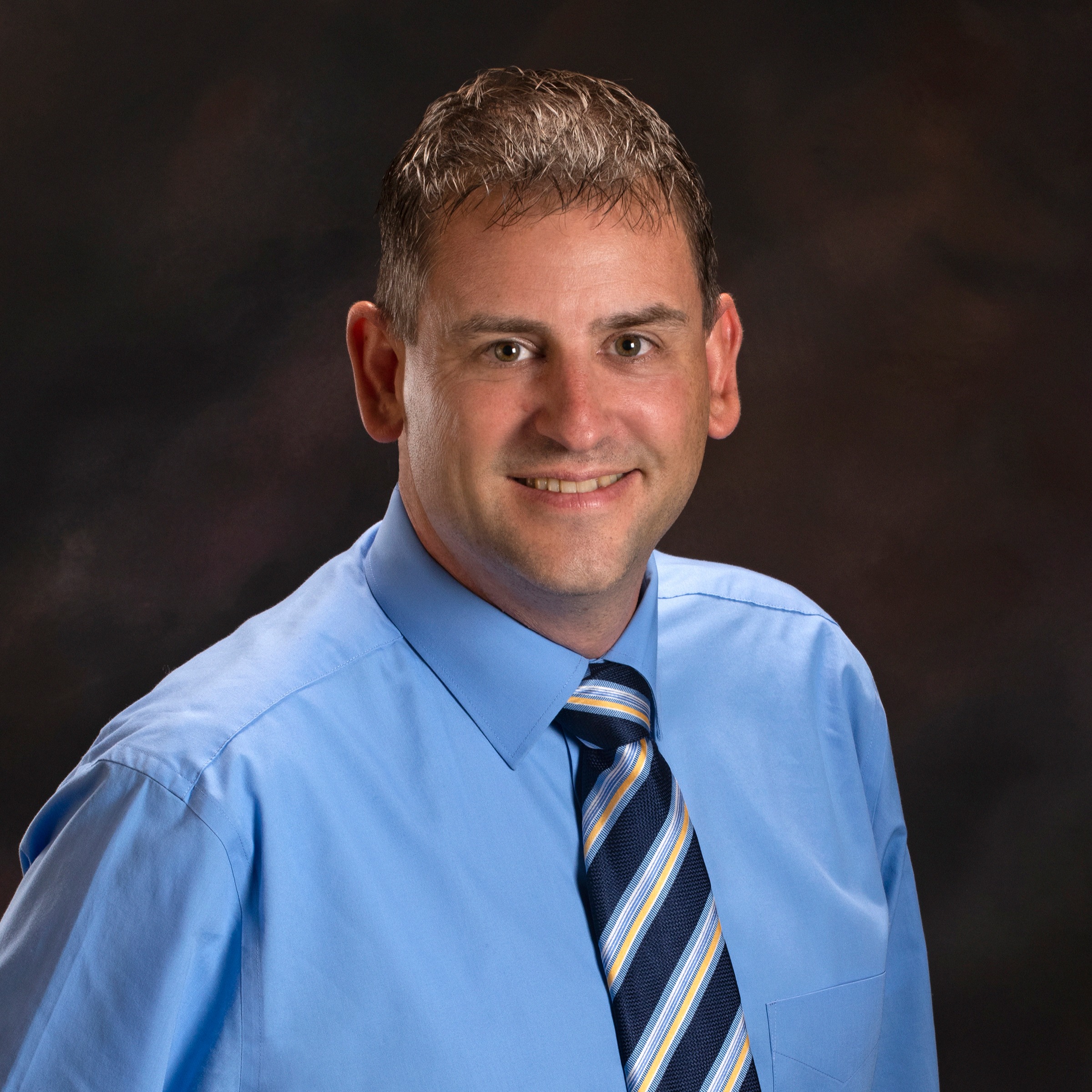 Person in light blue shirt and striped tie standing against a dark background, smiling.