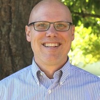 A bald man with glasses smiles in front of a tree. He wears a blue shirt with vertical stripes.