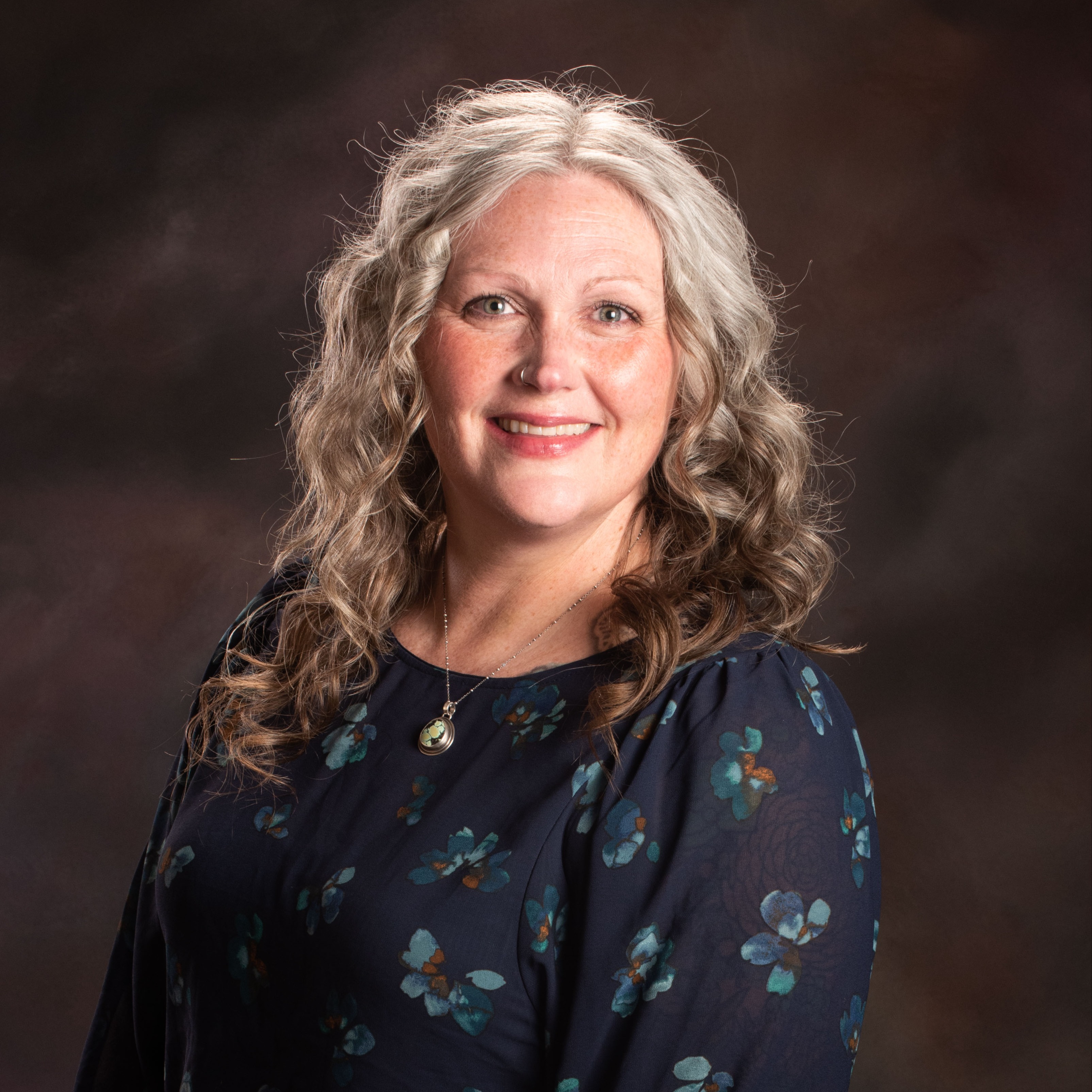 Woman with blonde curls wearing a dark blue blouse with floral patterns smiles at the camera.