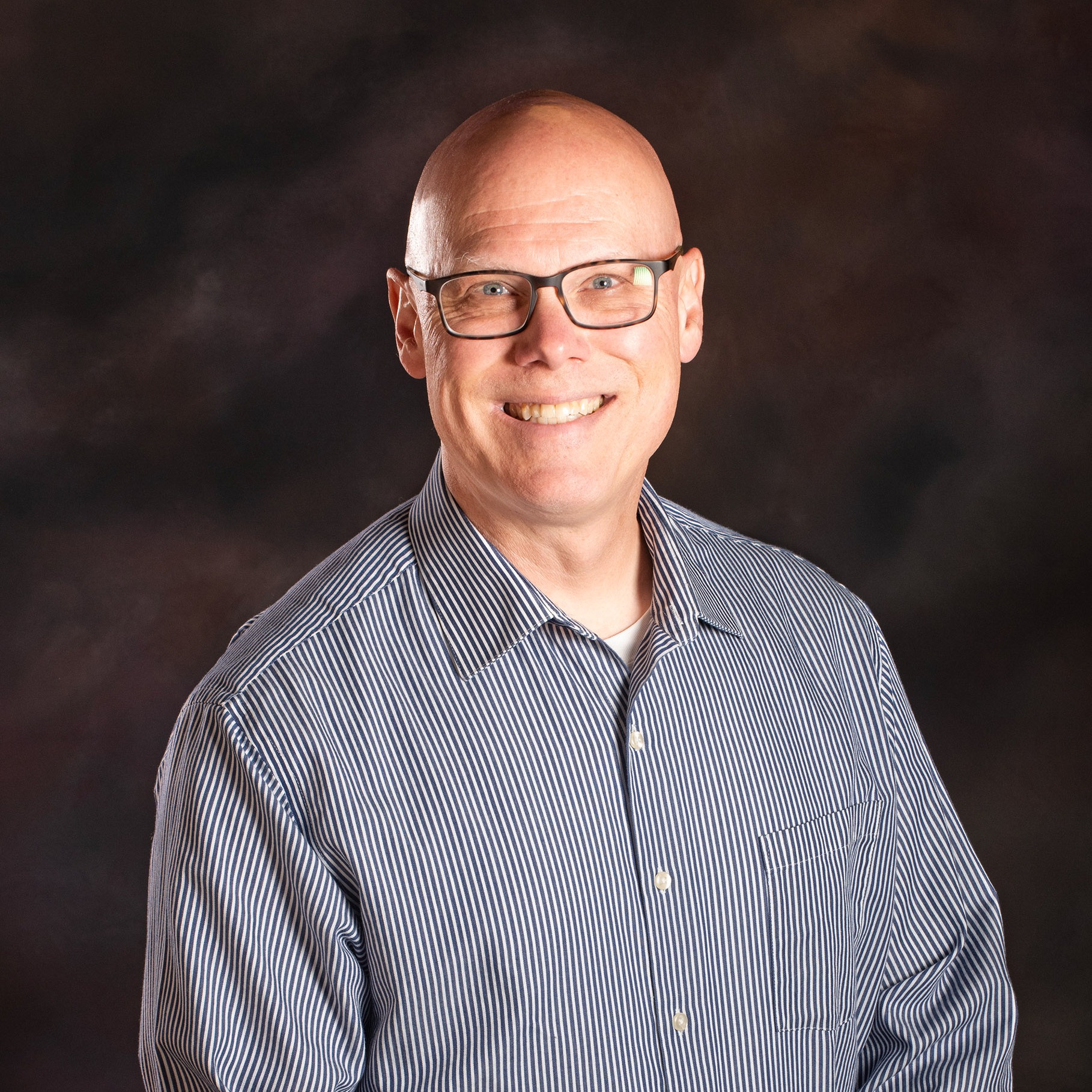 Man smiling with arms folded in front of a dark background, wearing glasses and a striped shirt.
