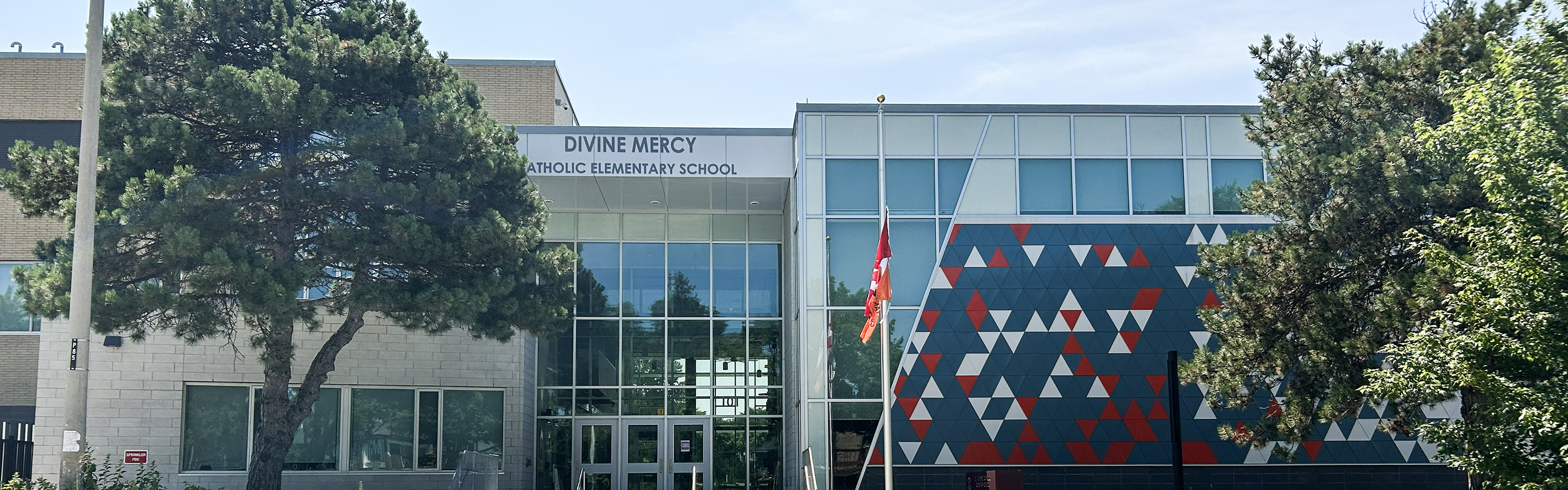Photo of the Divine Mercy Catholic School building, showing a modern building with portions of it in beige bricks, and the other as fully glass with a navy, red, and white triangle mosaic, nestled between trees and looking out onto the road. 