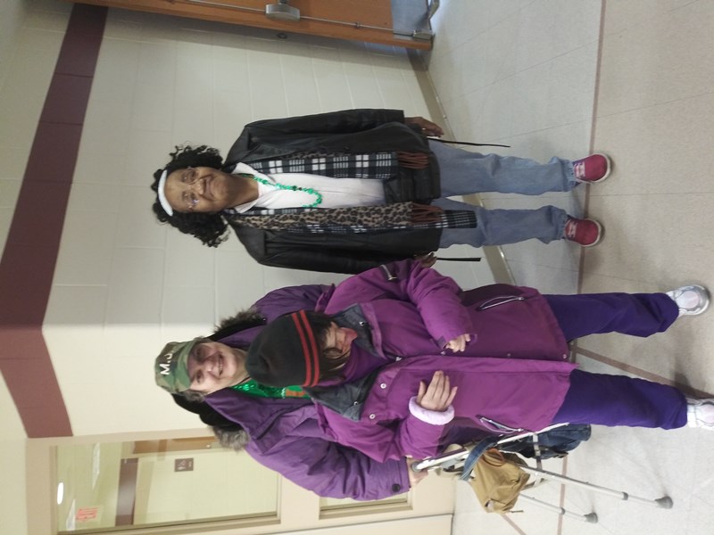 Three individuals are standing together in a hallway, dressed warmly in colorful winter clothing, with one person using a walker.