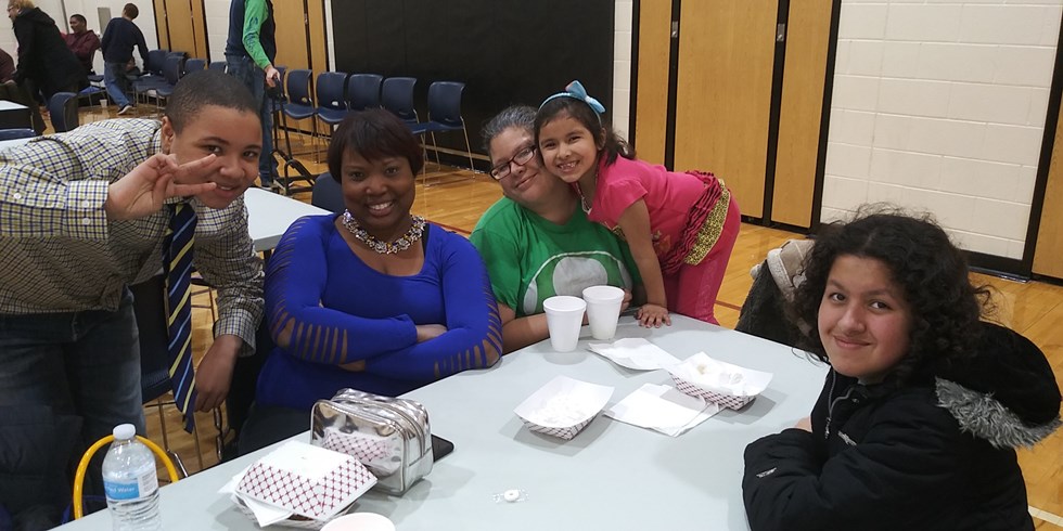 A group of five people, including children and adults, are smiling and posing together at a table with food containers in a community setting.
